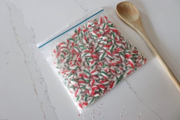 Freezer bag with crushed candy canes in it resting on a counter top with a wooden spoon.
