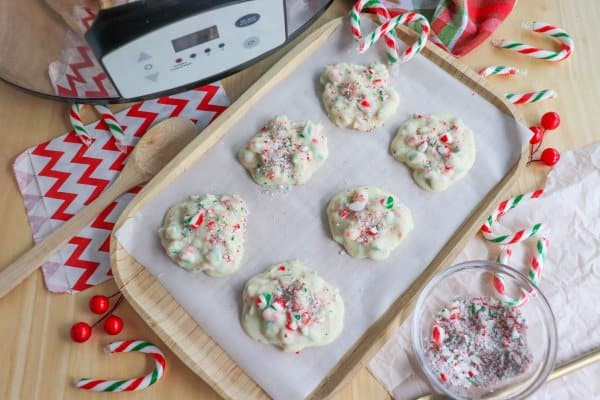 White chocolate clusters with candy cane sprinkles on a wood tray lined with parchment paper with a crockpot in the background.