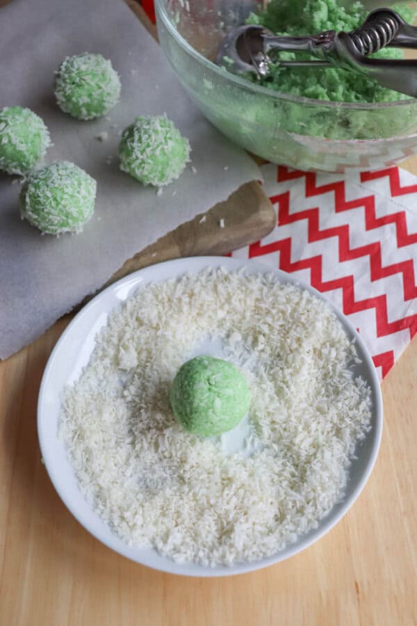 A green coconut ball on a white plate filled with shredded coconut resembles a tropical twist on the thin mint cookie recipe. More coconut balls and a scoop of green mixture are in the background, next to a red and white zigzag patterned cloth partially visible on the side.
