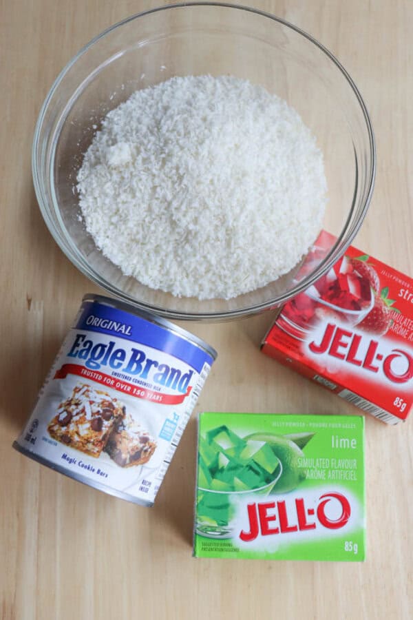 A glass bowl filled with shredded coconut sits on a wooden surface, surrounded by a can of Eagle Brand condensed milk, a box of strawberry Jell-O, and a box of lime Jell-O—perfect ingredients to experiment with your thin mint cookie recipe.