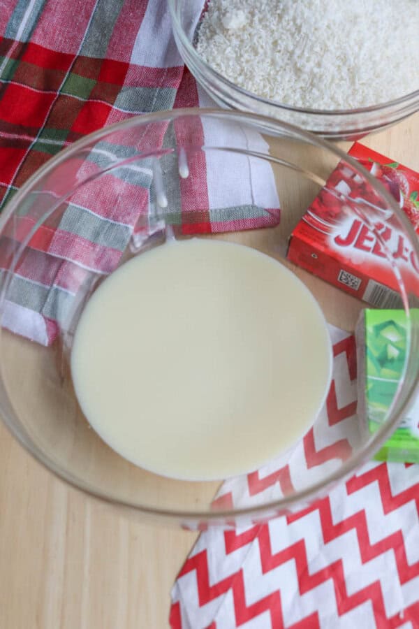 A bowl of liquid sits on a wooden table near a red and green checkered cloth, ready to inspire your next thin mint cookie recipe. Behind it are bowls of shredded coconut and boxes of flavored gelatin mix. A red and white zigzag patterned napkin is partially visible.