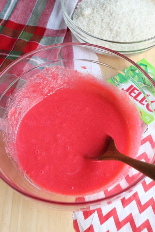 A clear glass bowl holds a bright red liquid mixture with a wooden spoon. Nearby, a packet of lime Jell-O hints at more flavors, while a bowl of white powder sits in the background, reminiscent of starting your favorite thin mint cookie recipe. A red-green plaid cloth and red chevron cloth complete the scene.