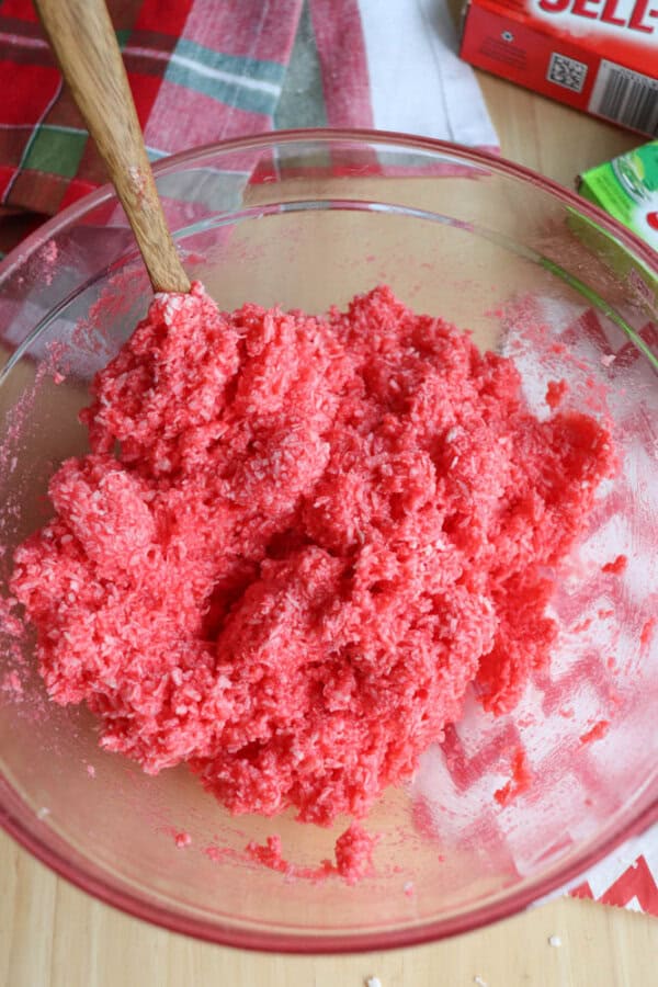 A clear glass bowl brims with a mixture of red, shredded coconut. A wooden spoon dips into the blend, evoking the richness of a thin mint cookie recipe. In the background, a red and green checkered cloth adds cheer next to a box of Jell-O.
