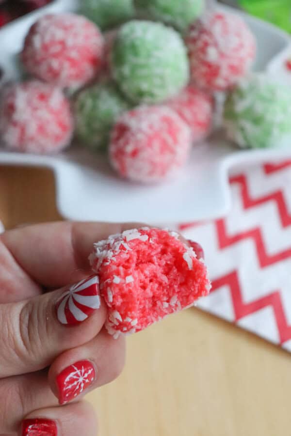 A person holding a pink and white candy coated in shredded coconut, with a bite taken out, savors the sweetness reminiscent of a thin mint cookie recipe. In the background, a plate of green and pink candies is visible. The person's nails are painted with festive red, white, and snowflake designs.