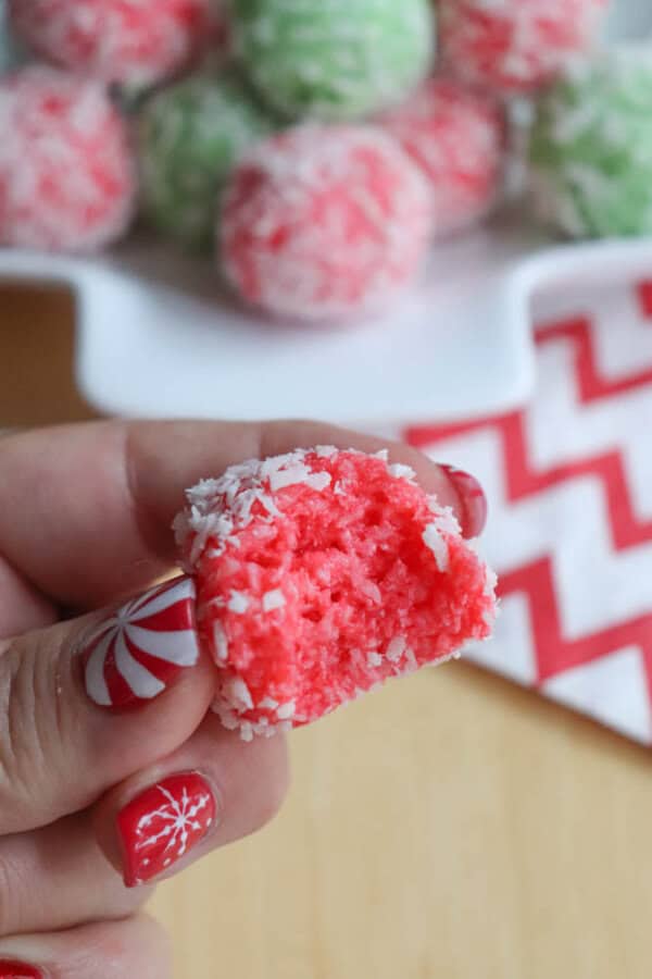A hand holds a bitten red coconut ball with a white coconut coating, reminiscent of a festive thin mint cookie. The person flaunts red and white nail art. In the background, a plate brims with more coconut balls in vibrant red and green hues, resting on a chevron-patterned cloth.