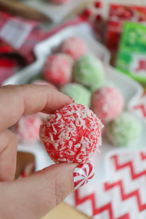 A hand holding a red and white coconut-covered candy ball evokes the holiday spirit. In the background, more candy balls in red, green, and white sit on a star-shaped plate, reminiscent of a festive thin mint cookie recipe. The person's nail is painted with a peppermint candy design.