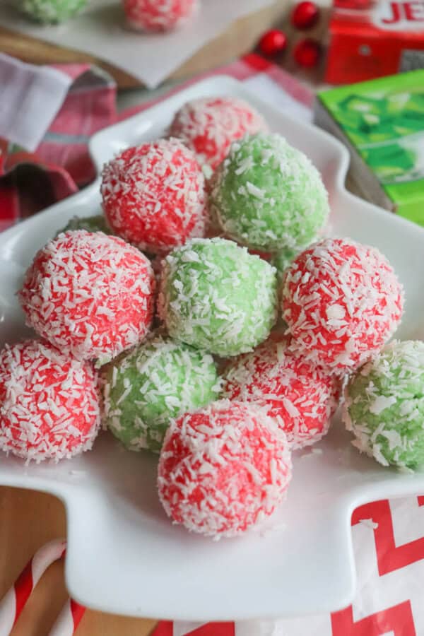 A white platter holds festive red and green balls covered in shredded coconut, reminiscent of a thin mint cookie recipe. The table is adorned with holiday-themed items in the background, including red ornaments and plaid fabric.