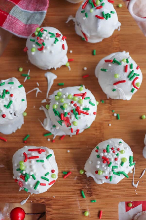 Festive cake balls coated in white icing, topped with red, green, and white sprinkles, arranged on a wooden surface. Reminiscent of rocky road crockpot candy, they're decorated with round sugar pearls and colorful sprinkles to evoke a warm holiday theme.