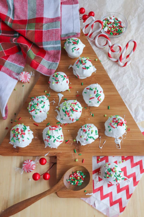 A wooden board displays rocky road crockpot candy coated in white chocolate, adorned with red, green, and white sprinkles. A plaid fabric, candy canes, peppermint candies, and a bowl of sprinkles surround the board, alongside chevron-patterned napkins.