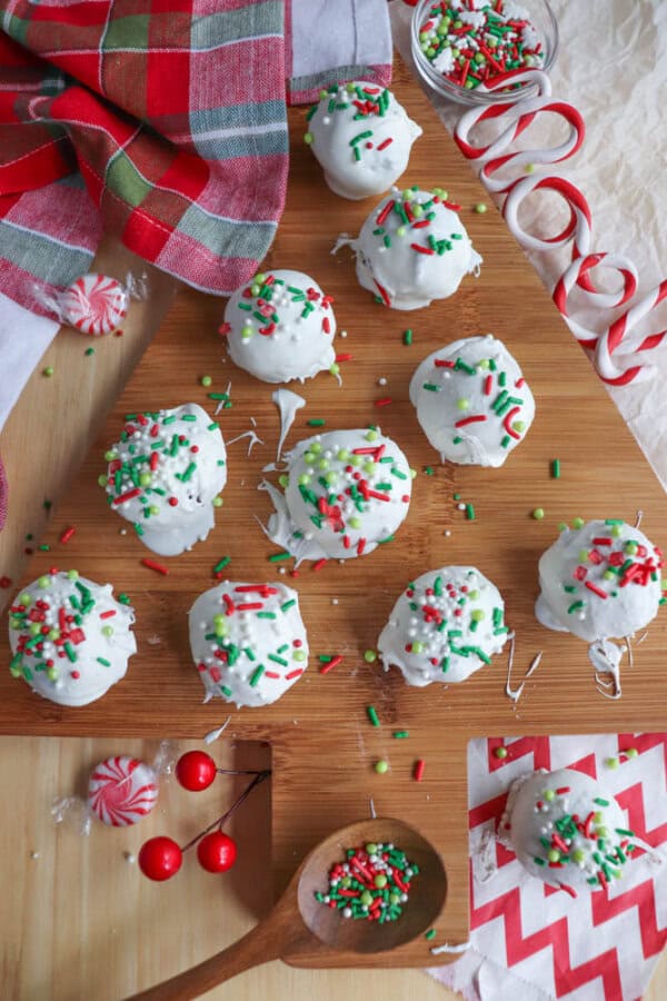 A wooden board displays white chocolate-covered rocky road treats topped with festive red, green, and white sprinkles. Nearby, there's a red and green plaid cloth, candy canes, red berries, and peppermint candies, creating a holiday theme. Perfectly easy to make in your crockpot!