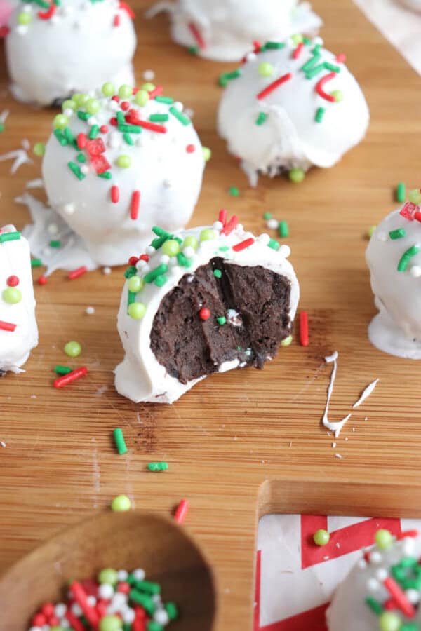Chocolate cake balls, reminiscent of rocky road treats, are coated in white chocolate and decorated with red, green, and white sprinkles. They sit on a wooden board beside a small spoon holding extra sprinkles. One ball is cut in half, revealing the decadent dark interior.