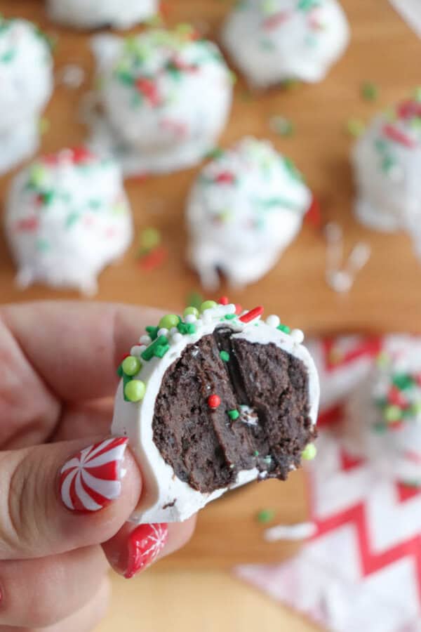 A close-up of a hand holding a chocolate truffle with white coating and festive green and red sprinkles resembles the playful look of rocky road candies. The truffle is bitten to reveal a dark, rich interior. More truffles are blurred in the background on a wooden surface.