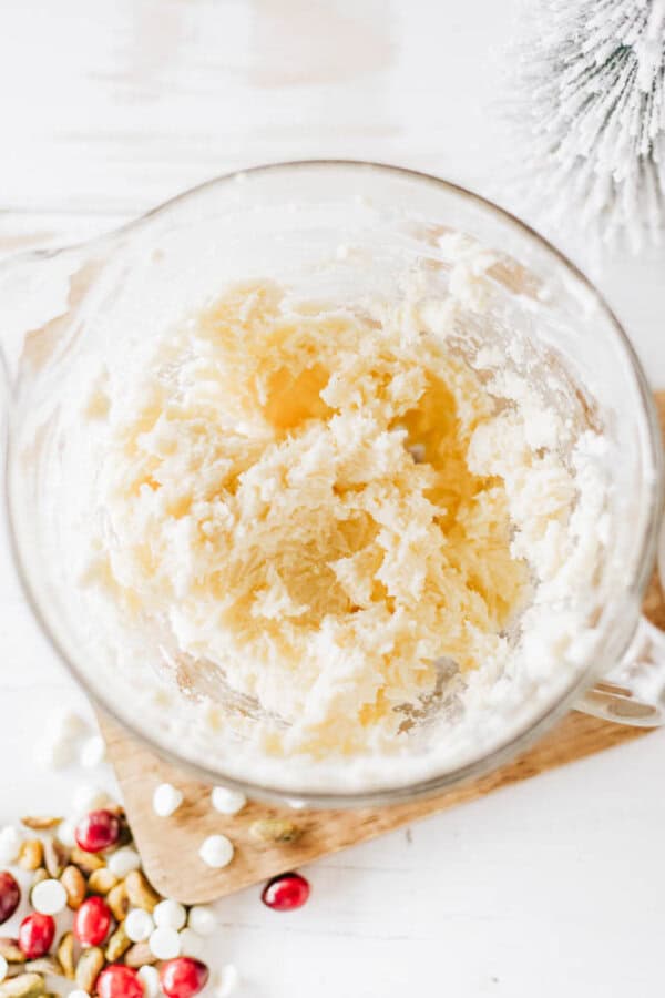 A glass bowl filled with creamy, textured cookie dough sits on a wooden board, reminiscent of rocky road. Scattered red, white, and green candy pieces add a festive touch, while a small frosted evergreen branch decorates the background.