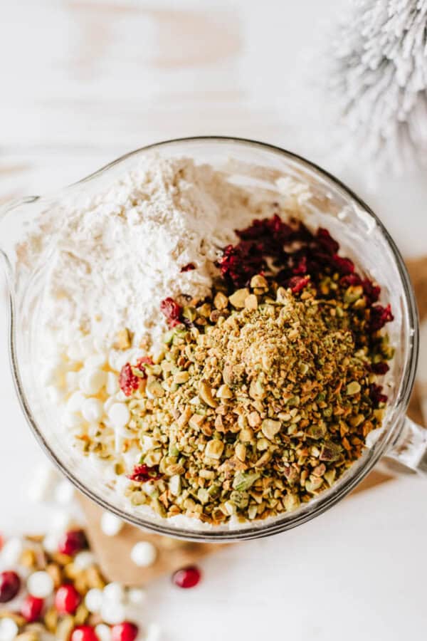 A glass bowl on a white surface contains flour, crushed pistachios, dried cranberries, and white chocolate chips—the essentials for a rocky road candy delight. A few ingredients are scattered around the bowl. A blurred, frosty decoration is visible in the background.