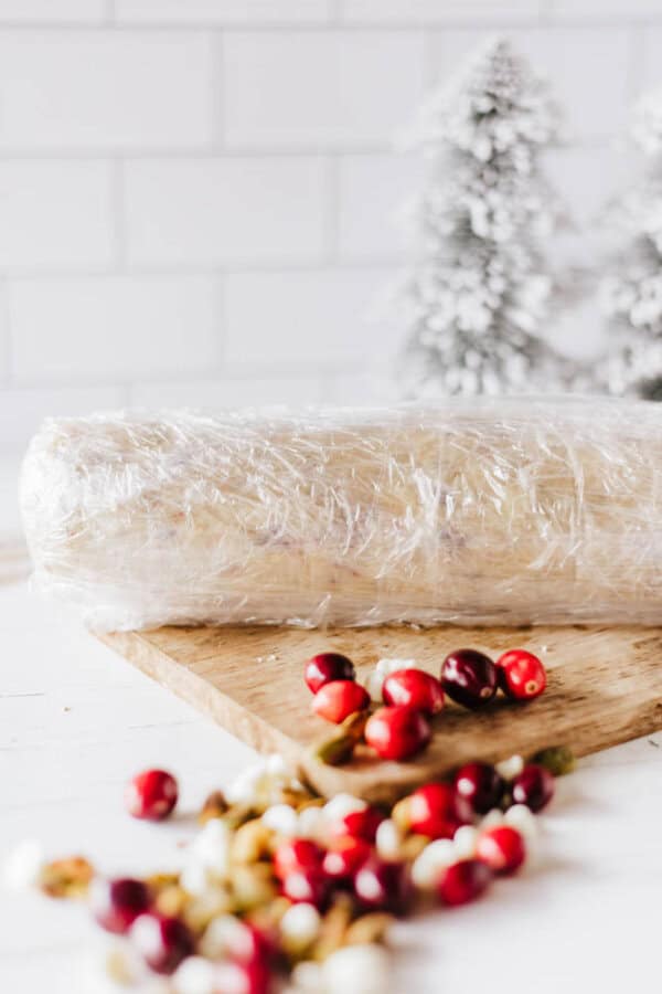 A log of dough wrapped in plastic sits on a wooden board, evoking the look of homemade rocky road crockpot candy. A few cranberries and nuts are scattered nearby, adding to the festive touch. In the background, snowy evergreen trees are blurred, completing the holiday setting.