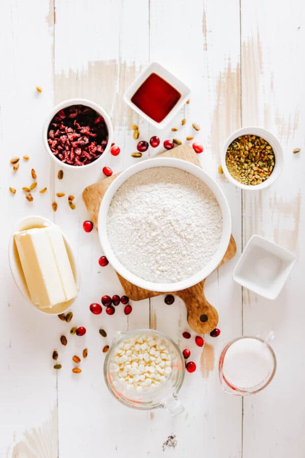 Top-down view of baking ingredients on a white wooden surface: a bowl of flour, butter sticks, cranberries, pistachios, white chocolate chips, sugar, salt, and a small bowl of vanilla extract. Perfect for crafting rocky road crockpot candy with scattered cranberries and nuts adding charm.