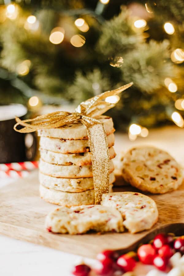 A festive stack of cookies tied with a gold ribbon is placed on a wooden board, reminiscent of rocky road crockpot candy. The cookies boast visible cranberry and nut pieces. In the background, a softly focused Christmas tree glows with warm lights, adding to the cozy holiday atmosphere.