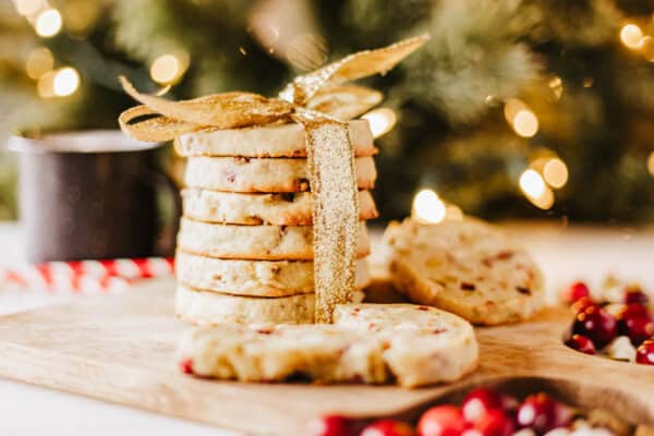 A stack of rocky road cookies tied with a gold ribbon sits on a wooden board. The cookies are speckled with cranberries and nuts. A few are broken in front, with scattered cranberries around. A blurred festive background with lights enhances this sweet scene.