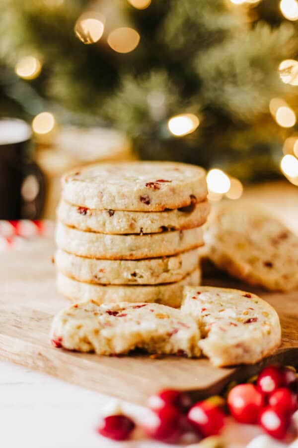 A stack of cranberry cookies on a wooden board, with one broken cookie and bits of rocky road in the foreground. Scattered cranberries and blurred festive lights in the background create a warm, holiday atmosphere.