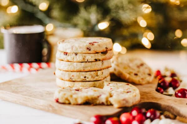 A stack of cranberry cookies on a wooden board, with more cookies scattered around, evokes the delightful mess of rocky road crockpot candy. Fresh cranberries are scattered nearby, while a blurred background with festive lights and a mug adds to the cozy atmosphere.