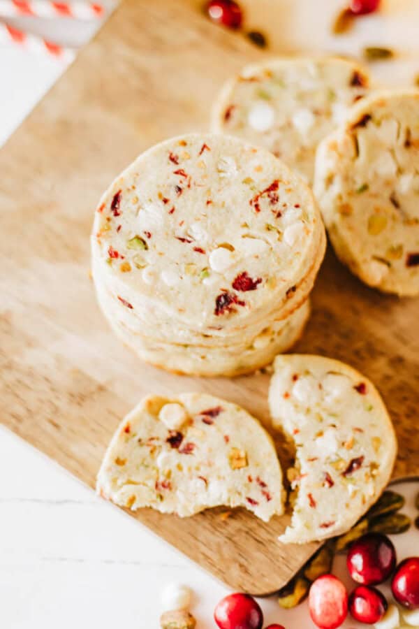 A stack of sliced holiday cookies on a wooden board, featuring cranberries and nuts, evokes flavors reminiscent of rocky road crockpot candy. Some cookies are slightly broken, with scattered cranberries adding an inviting touch nearby.