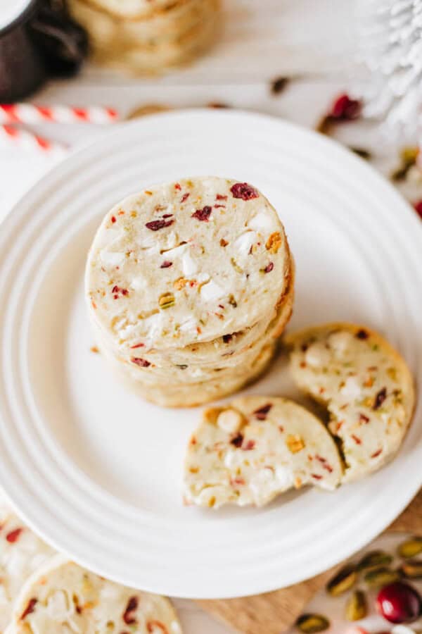 A stack of cranberry pistachio cookies is arranged on a white plate, resembling the decadent mix found in rocky road crockpot candy. One cookie is broken in half, showcasing its crumbly texture. The cookies are speckled with red cranberries and green pistachios, surrounded by festive decor.