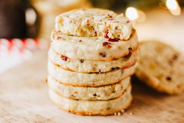 A stack of round cookies with cranberries and nuts sits on a wooden surface, reminiscent of a homemade rocky road treat. The top cookie has a bite taken out of it. Soft, warm lighting in the background creates a cozy atmosphere.