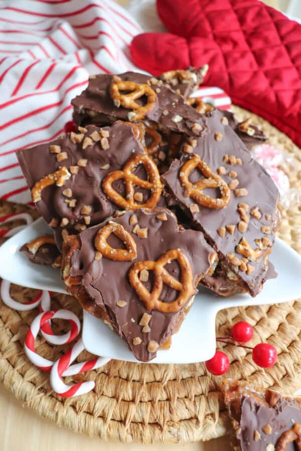 A plate of chocolate toffee bark topped with pretzels and toffee pieces, reminiscent of rocky road indulgence. The bark is stacked on a woven placemat, surrounded by red oven mitts, striped cloth, candy canes, and red candies creating a cozy crockpot candy vibe.