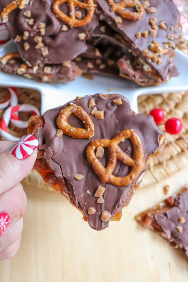 A person holds a piece of chocolate bark, reminiscent of rocky road treats, topped with pretzels and toffee bits. More pieces are stacked in the background. The person's nails are painted with festive red and white designs, adding to the cozy vibe of enjoying crockpot candy delights.