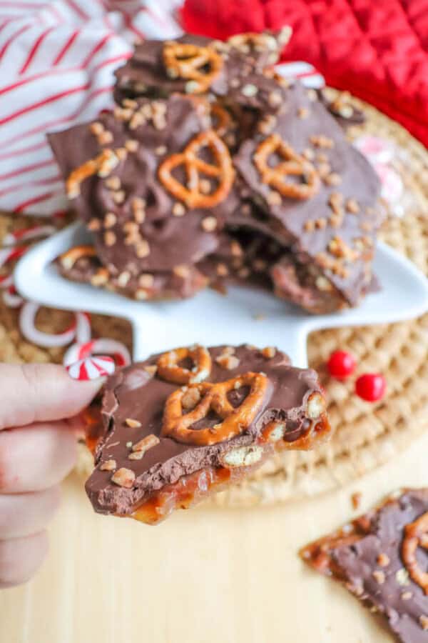 A person is holding a piece of rocky road chocolate bark topped with pretzels and nuts. More pieces are piled on a white plate in the background, next to a red and white striped cloth. Peppermint candies and red berries are also visible.