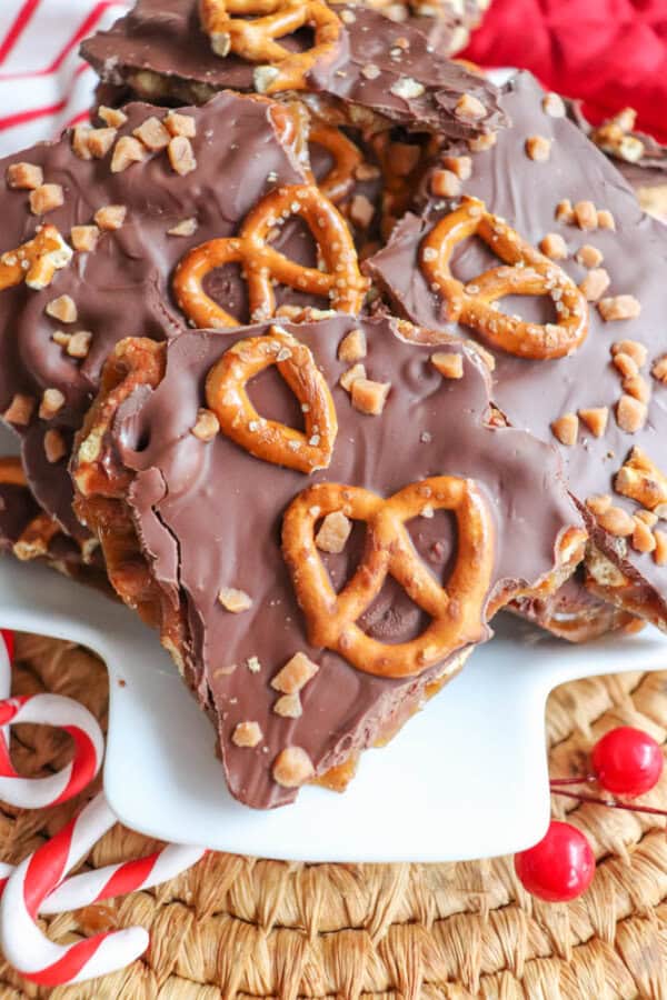 A plate of chocolate bark topped with pretzels and toffee bits sits invitingly. Resembling rocky road crockpot candy, the treats are artfully arranged on a white plate, encircled by candy canes and red berries on a woven mat.
