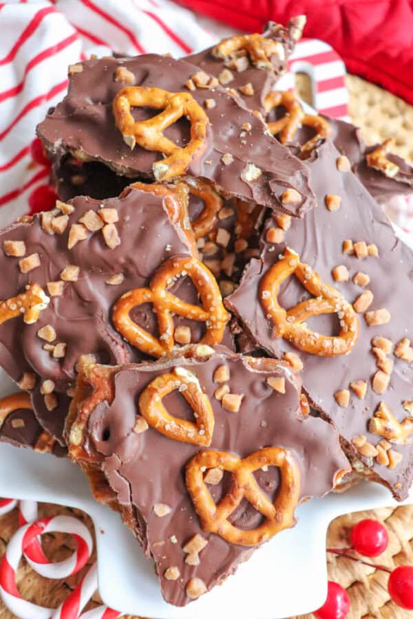 A plate of chocolate bark reminiscent of rocky road, topped with crushed toffee and whole pretzels. The bark is broken into uneven pieces, evoking a rustic charm. In the background, a red and white striped cloth adds a cozy touch.