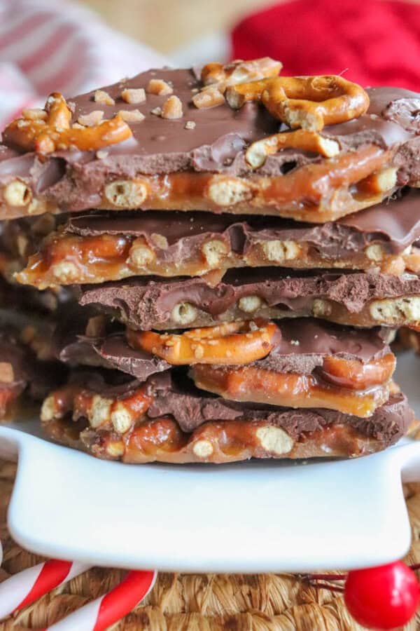 A stack of rocky road chocolate bark with pretzels and caramel is piled on a white dish. The bark boasts a glossy chocolate coating, interspersed with crunchy pretzels and gooey caramel. Candy canes are visible at the bottom of the image.