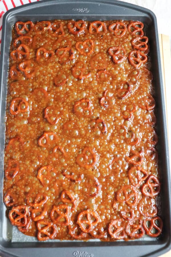 A baking sheet filled with a caramel-like mixture, reminiscent of rocky road, is topped with pretzels. The concoction bubbles enticingly as the pretzels are evenly spread across the surface. The sheet rests invitingly on a light background, tempting anyone nearby to sample its sweetness.