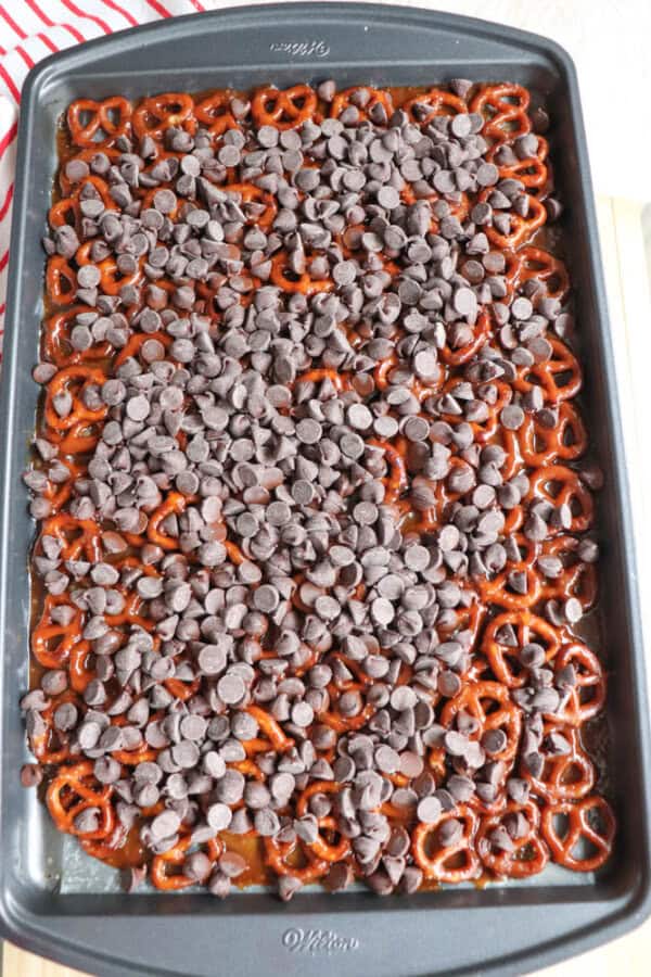 A baking tray filled with a layer of pretzels topped with chocolate chips resembles a rocky road treat. The pretzels are arranged evenly, and the chocolate chips cover them generously. The tray sits on a countertop, with a striped cloth partially visible in the background.