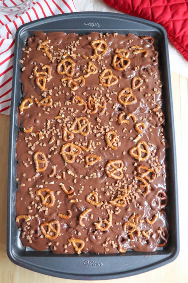 A baking tray filled with decadent rocky road dessert, topped with pretzels and small crushed candy pieces. A red oven mitt and a striped cloth are partially visible beside the tray.