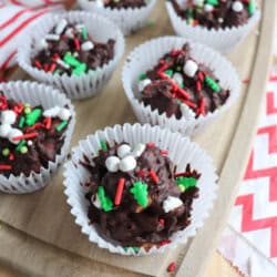 Rocky road crockpot candy treats decorated with sprinkles, mini marshmallows, and Christmas-themed toppings in white paper cups are arranged on a wooden tray. A red and white striped cloth is underneath the tray.