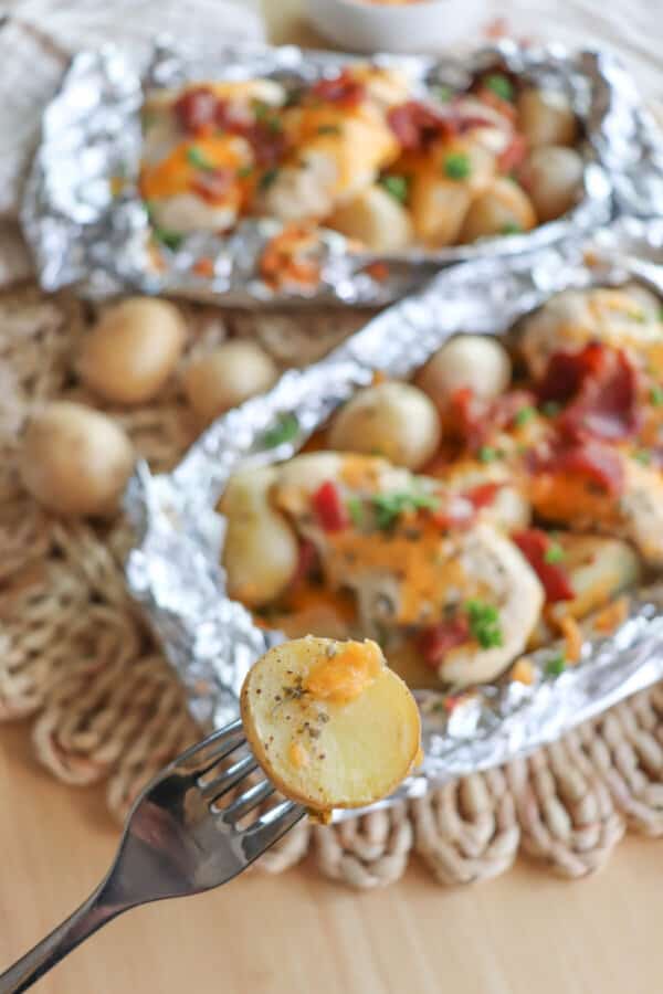 A fork with a baby potato over chicken and baby potatoes covered in ranch seasoning, bacon bits and melted cheese in a foil packet.