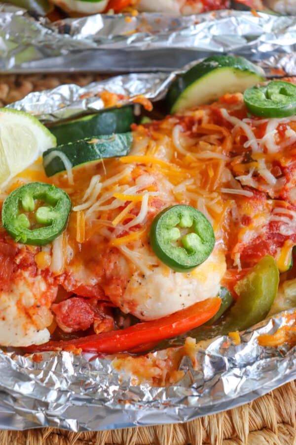 Close up of chicken on a bed of sliced bell peppers and zucchini topped with melted shredded cheese and jalapeno slices with a lime wedge garnish in a foil packet.