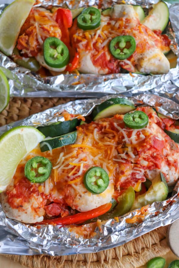 Chicken on a bed of sliced bell peppers and zucchini topped with melted shredded cheese and jalapeno slices with a lime wedge garnish in a foil packet.