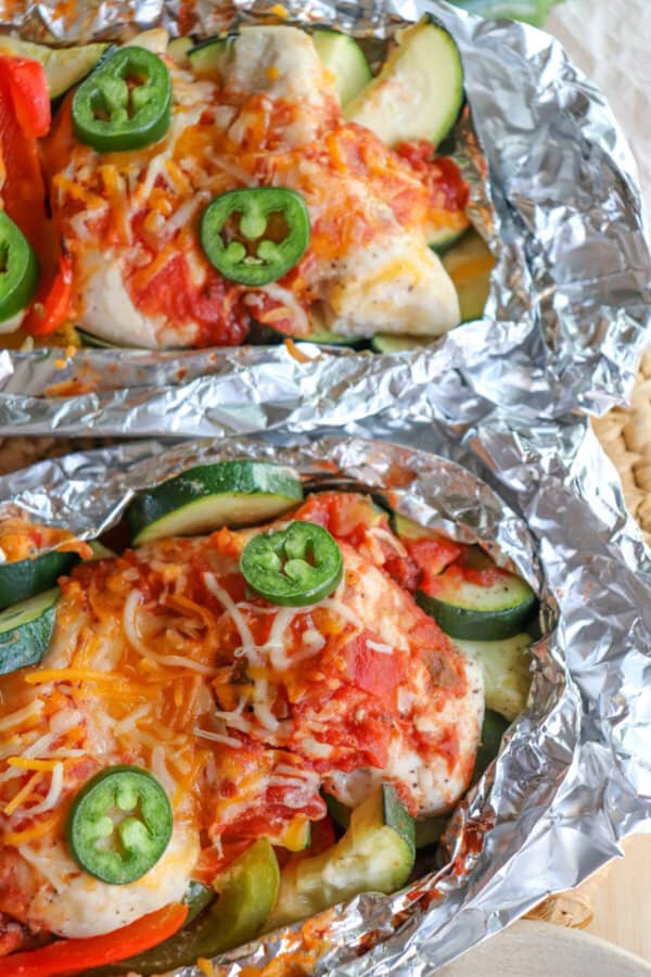 Chicken on a bed of sliced bell peppers and zucchini topped with melted shredded cheese and jalapeno slices with a lime wedge garnish in a foil packet.