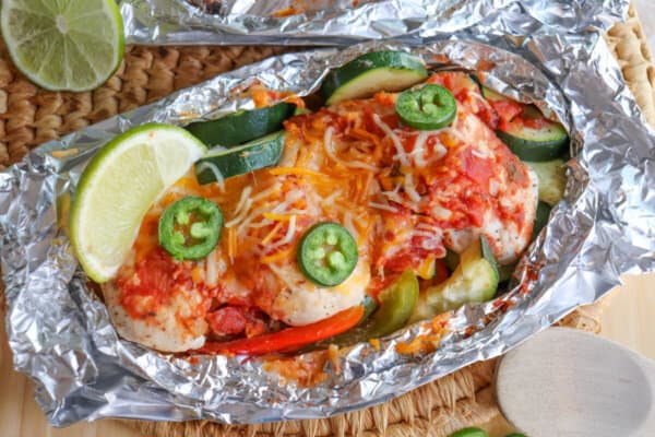 Chicken on a bed of sliced bell peppers and zucchini topped with melted shredded cheese and jalapeno slices with a lime wedge garnish in a foil packet.