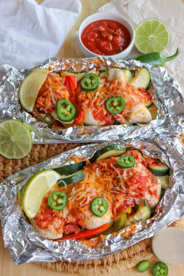 Chicken on a bed of sliced bell peppers and zucchini topped with melted shredded cheese and jalapeno slices with a lime wedge garnish in a foil packet.