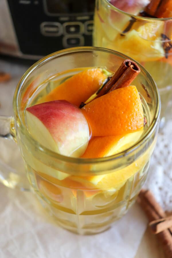 A clear glass of apple cider with chunks of orange, apple and a cinnamon stick for garnish.