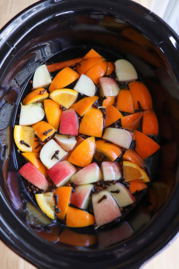 A black crockpot full of apple cider with apples, oranges, cinnamon sticks and cloves floating on top.