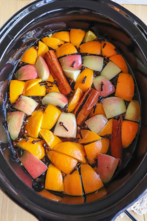 A black crockpot full of apple cider with apples, oranges, cinnamon sticks and cloves floating on top.