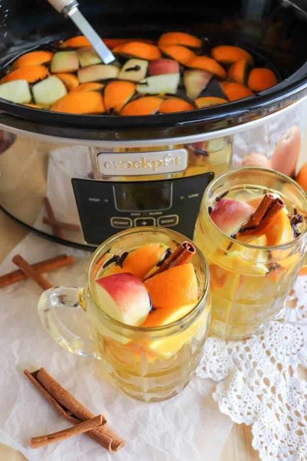 Apple cider in a black crockpot with two clear glasses full that have orange and apple slices for garnish.