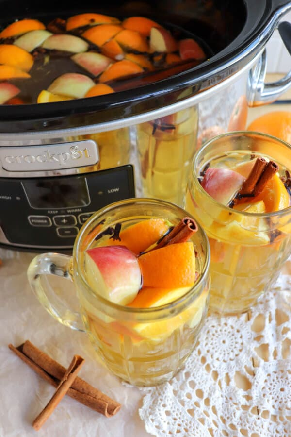 Apple cider in a black crockpot with two clear glasses full that have orange and apple slices for garnish.