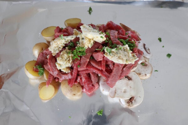 Raw steak, sliced mushrooms and baby potatoes topped with garlic butter and parsley on a sheet of foil.