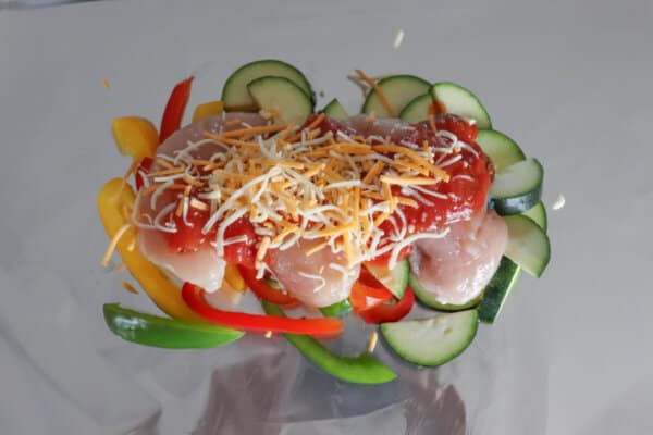 Raw chicken breast on a bed of vegetables topped with shredded cheese on a sheet of foil.
